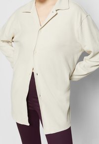 Camisa abotonada de color beige claro con cuello, hecha de material suave. Presenta un corte relajado y botones visibles en la parte delantera.