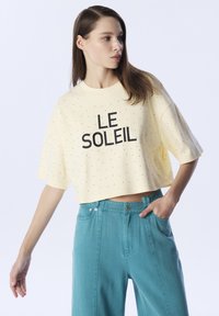 Gelbes, kurzes T-Shirt mit schwarzem "LE SOLEIL"-Text und kleinen Perforationen. Kombiniert mit hoch taillierten türkisfarbenen Hosen mit Knopf und Taschen.