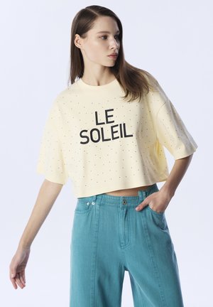 Gelbes, kurzes T-Shirt mit schwarzem "LE SOLEIL"-Text und kleinen Perforationen. Kombiniert mit hoch taillierten türkisfarbenen Hosen mit Knopf und Taschen.