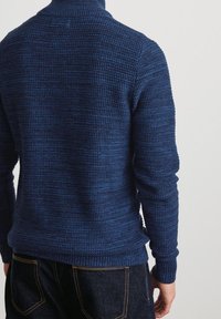 Marineblauer Strickpullover mit geripptem Kragen und Bündchen. Verfügt über ein strukturiertes Muster, eine enganliegende Passform und einen leicht längeren Saum hinten. Dunkelblaue Jeans sind sichtbar.