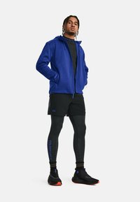 Sportliches Outfit, bestehend aus einem blauen Reißverschluss-Hoodie, schwarzen Shorts und schwarzen Leggings mit blauen Akzenten. Schwarze Turnschuhe mit orangefarbenen Sohlen.