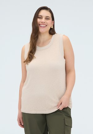 Smilende kvinde med langt brunt hår iført en beige ærmeløs top og olivengrønne cargobukser mod en lys baggrund.