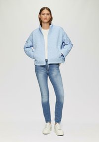 Chaqueta acolchada de un azul claro con cuello alto, bolsillos laterales y dobladillo con cordón, combinada con jeans ajustados azules y zapatillas blancas.