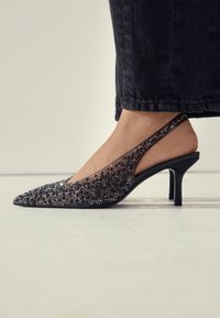 Crna cipela s remenom oko peta ukrašena sjajnim ukrasima. Šiljasti vrh, prozirni bočni dijelovi i tanak potpetica na štiklu.