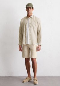 Beige linnen overhemd met knopen, witte T-shirt, beige shorts, groene pet en tan schoenen. Eenvoudig ontwerp met zachte texturen en neutrale kleuren.