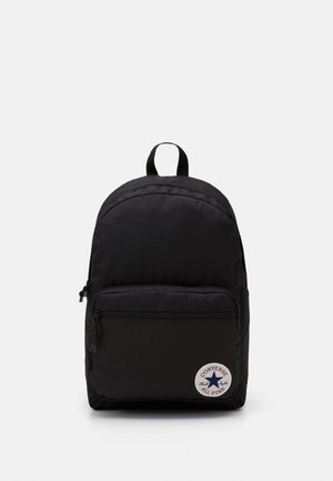 Sac à dos noir avec poche avant zippée et patch rond blanc avec le logo Converse All Star dans le coin inférieur droit.