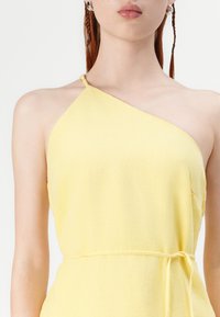 Asymmetrical one-shoulder yellow dress made of lightweight fabric, with a textured finish and a tied waistband for a defined shape.