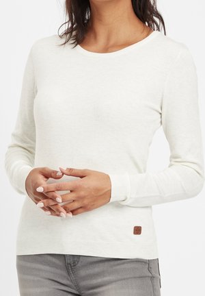 Femme portant un pull blanc à manches longues et un pantalon gris, les mains doucement croisées à la taille, avec un petit écusson brun sur l'ourlet du pull.