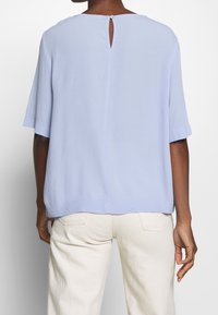 Blusa azzurra chiara a maniche corte con apertura a forma di chiave sul retro, texture morbida e taglio dritto. Si abbina a pantaloni di colore chiaro.