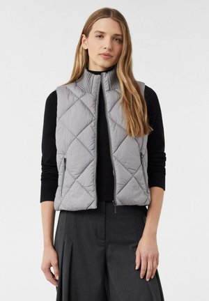 Graue, gesteppten Pufferweste mit hohem Kragen, die über einem schwarzen Pullover getragen wird. Sie hat einen Frontreißverschluss und Seitentaschen. Glatte Textur.