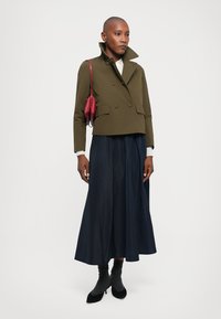 Jachetă verde măslinie cu două rânduri de nasturi, scurtă, cu guler înalt, asortată cu o fustă lungă de culoare bleumarin. Accesoriile includ o geantă roșie și tocuri negre.