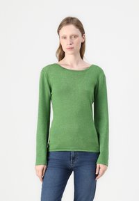 Grønn langermet genser med avslappet båtutringning og en glitrende tekstur. Kombinert med tettsittende blå jeans.