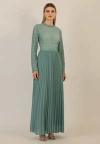 Lungo vestito teal con corpetto in pizzo e gonna plissettata. Presenta maniche lunghe e un collo a scallop per un tocco di dettaglio in più.