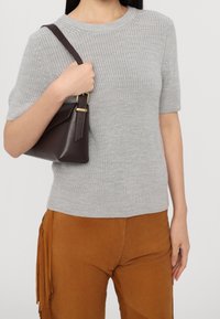 Pull gris à manches courtes en tricot texturé associé à un pantalon en suède beige avec des accents à franges. Un sac à main marron foncé est porté à l'épaule.