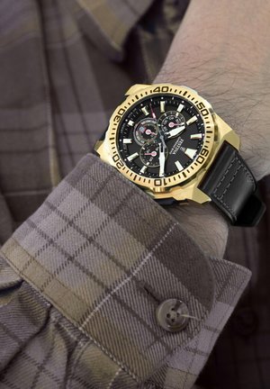 Orologio da polso dorato con quadrante nero, accenti in argento, funzioni cronografiche e cinturino in pelle nera, esposto su una manica di camicia scozzese.