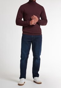 Pull à col roulé bordeaux, en tissu extensible, au design ajusté ; associé à un jean bleu foncé et des baskets blanches avec semelles beige.