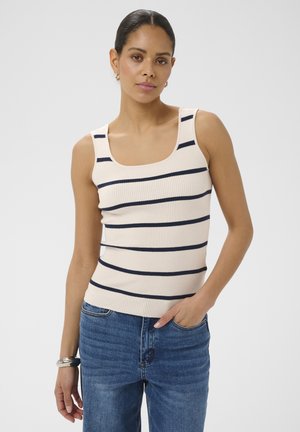 Femme aux cheveux foncés portant un débardeur blanc côtelé sans manches avec des rayures horizontales noires et un jean bleu, debout avec une main dans la poche.