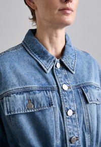 Világoskék farmerdzseki, kollárral, ezüst színű fém gombokkal, két mellzsebbel és látható varrási részletekkel.