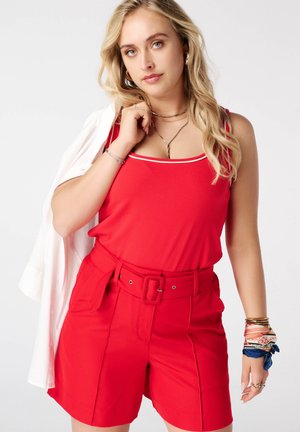 Blonde vrouw met een rode mouwloze top en hooggetailleerde rode short met riem, die een wit jasje over haar schouder houdt.