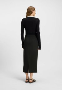 Haut noir à manches longues avec un col blanc, associé à une jupe midi verte foncée avec une fente arrière, portée avec des sandales plates noires.