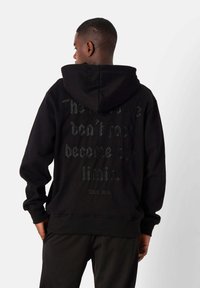 Sudadera negra con capucha grande, que presenta un texto en relieve texturizado en la parte posterior. Puños y dobladillo acanalados, fabricada con una tela suave.
