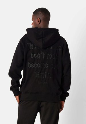 Sudadera negra con capucha grande, que presenta un texto en relieve texturizado en la parte posterior. Puños y dobladillo acanalados, fabricada con una tela suave.