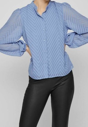 Blå gennemsigtig bluse med chevronmønster og puffede ærmer, sat sammen med sorte, tætsiddende bukser, modelens hænder hvilende på hoften.