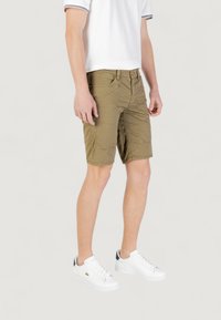 Pantaloni corti verde oliva con una texture leggera e liscia, dotati di numerose tasche e chiusura con zip, abbinati a sneakers bianche.