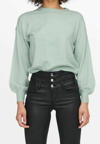 Mujer con un suéter de manga larga en verde menta metido en pantalones negros de cintura alta con tres botones plateados.