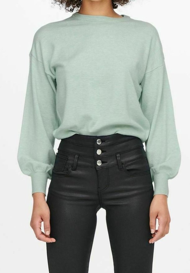 Mujer con un suéter de manga larga en verde menta metido en pantalones negros de cintura alta con tres botones plateados.