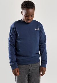 Sudadera azul marino hecha de mezcla de algodón, con cuello redondo, puños acanalados y un pequeño logo blanco en el pecho. Se combina con jeans grises.