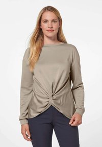 Beige Langarmoberteil mit Knotendetail an der Taille, aus weichem Stoff gefertigt. Mit Rundhalsausschnitt und lockerer Passform. Kombiniert mit dunklen Hosen.