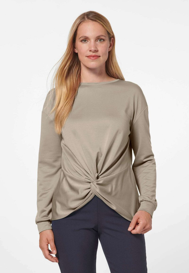 Beige Langarmoberteil mit Knotendetail an der Taille, aus weichem Stoff gefertigt. Mit Rundhalsausschnitt und lockerer Passform. Kombiniert mit dunklen Hosen.