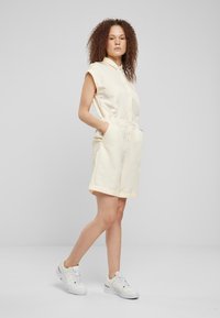 Lys gul kortermet romper med knapping foran, elastisk midje og side lommer. Brukt med hvite sneakers, med en glatt tekstur.
