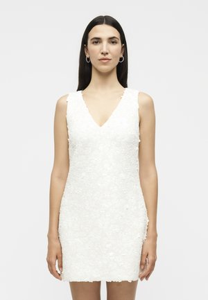 Γυναίκα με μακριά σκούρα μαλλιά που φορά κρίκους και λευκό υφασμάτινο μίνι φόρεμα χωρίς μανίκια, στέκεται μπροστά σε απλό φόντο.