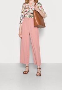 Kvinna iklädd rosa vida byxor, blommig blus, bruna lädersandaler och bär en stor brun läderhandväska.