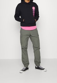 Svart hoodie med grafiskt tryck, rosa undertröja, olivfärgade cargo-byxor och svarta högskor med vita accenter. Avslappnad outfit.