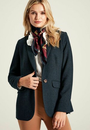 Dark green herringbone blazer with a single button closure and two front pockets, paired with a white ruffled blouse and patterned scarf.