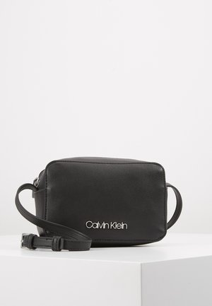 Czarna prostokątna skórzana torebka na ramię z regulowanym paskiem i srebrnym logo "Calvin Klein" z przodu, prezentowana na białej powierzchni.