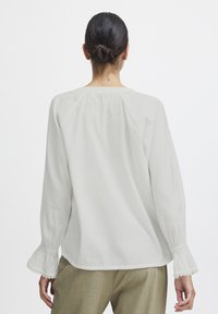 Blusa bianca a maniche lunghe realizzata in tessuto leggero. Presenta dettagli arricciati alle cuciture delle spalle e polsini con volant. Abbinata a pantaloni beige.