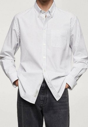 Man draagt een wit overhemd met dunne zwarte verticale strepen, handen in zijn zakken, gecombineerd met donkere spijkerbroek.