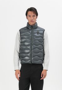 Dark gray puffer vest with a quilted wave pattern, high collar, and front zipper. Worn over a striped long-sleeve shirt, minimalistic design.