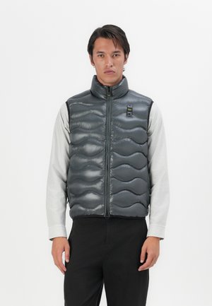 Chaleco acolchado gris oscuro con patrón de olas acolchadas, cuello alto y cremallera frontal. Llevado sobre una camiseta de rayas de manga larga, diseño minimalista.