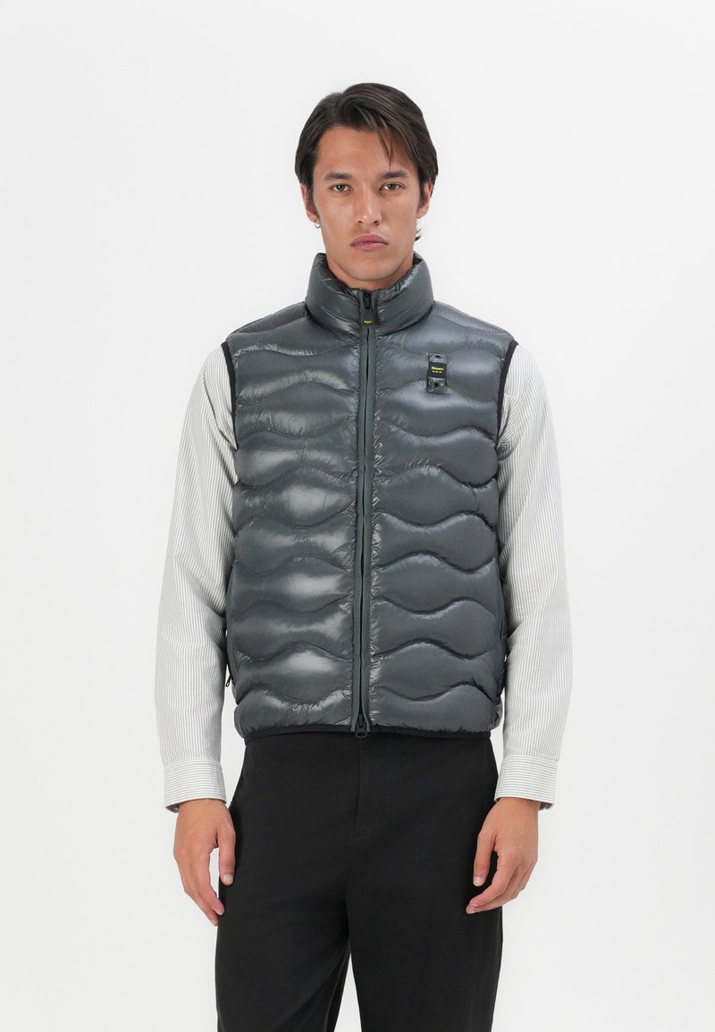 Dark gray puffer vest with a quilted wave pattern, high collar, and front zipper. Worn over a striped long-sleeve shirt, minimalistic design.