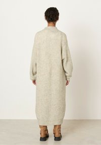 Svetlo bež, rebrasti pleteni dolg kardigan z velikimi rokavi, oblikovan s spuščeno ramo in zaobljenim robom. Tkanina se zdi mehka in teksturirana.