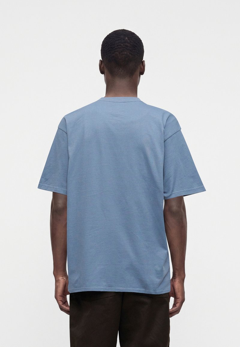 Uomo che indossa una t-shirt azzurro chiaro semplice a maniche corte e pantaloni scuri, visto di spalle su uno sfondo bianco uniforme.