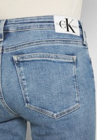Close-up of light blue denim jeans back pocket with visible Calvin Klein Jeans logo patch on waistband.
