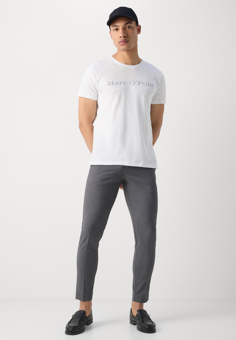 T-shirt bianca in cotone con lettering "Marc O'Polo", pantaloni grigi su misura e mocassini neri, indossati con un cappellino nero. Design minimal, forma aderente.