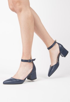 Woman's legs wearing navy blue leather ankle-strap block heels, one foot lifted, against white background.