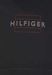 Sudadera negra con la palabra "HILFIGER" en letras blancas en negrita, contorneadas en rojo, sobre un fondo de textura de tela suave.